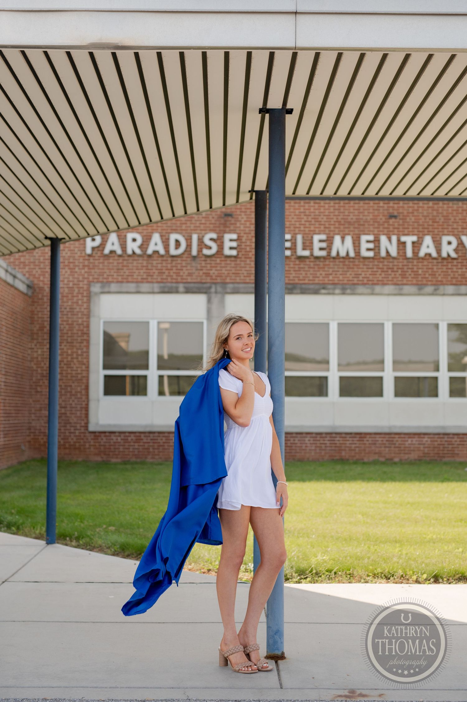 Graduate in blue cap and gown poses with a white dress against Paradise Elementary school building.