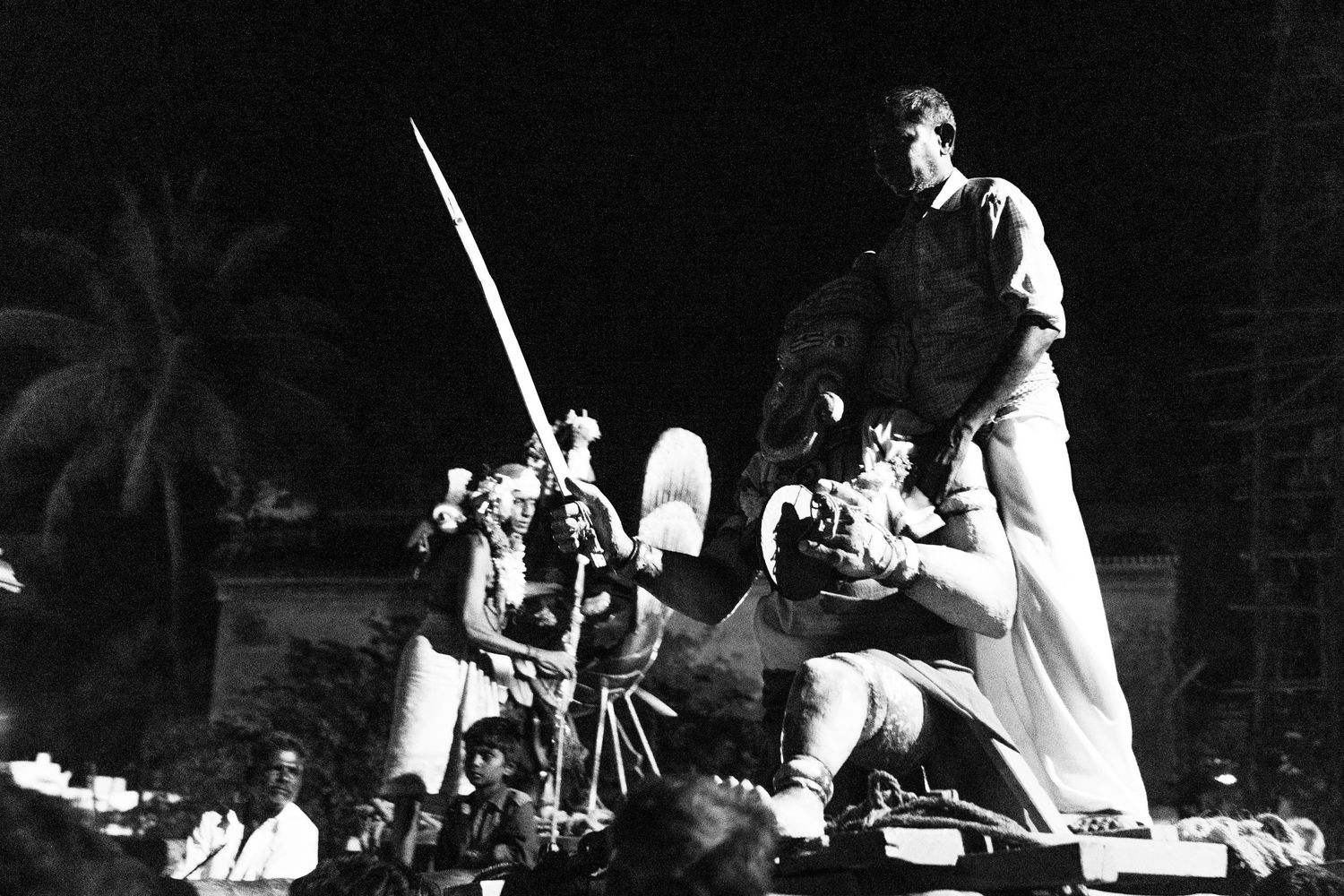 Traditional Hawaiian performers stand atop a stage with ceremonial spears during a nighttime cultural performance.