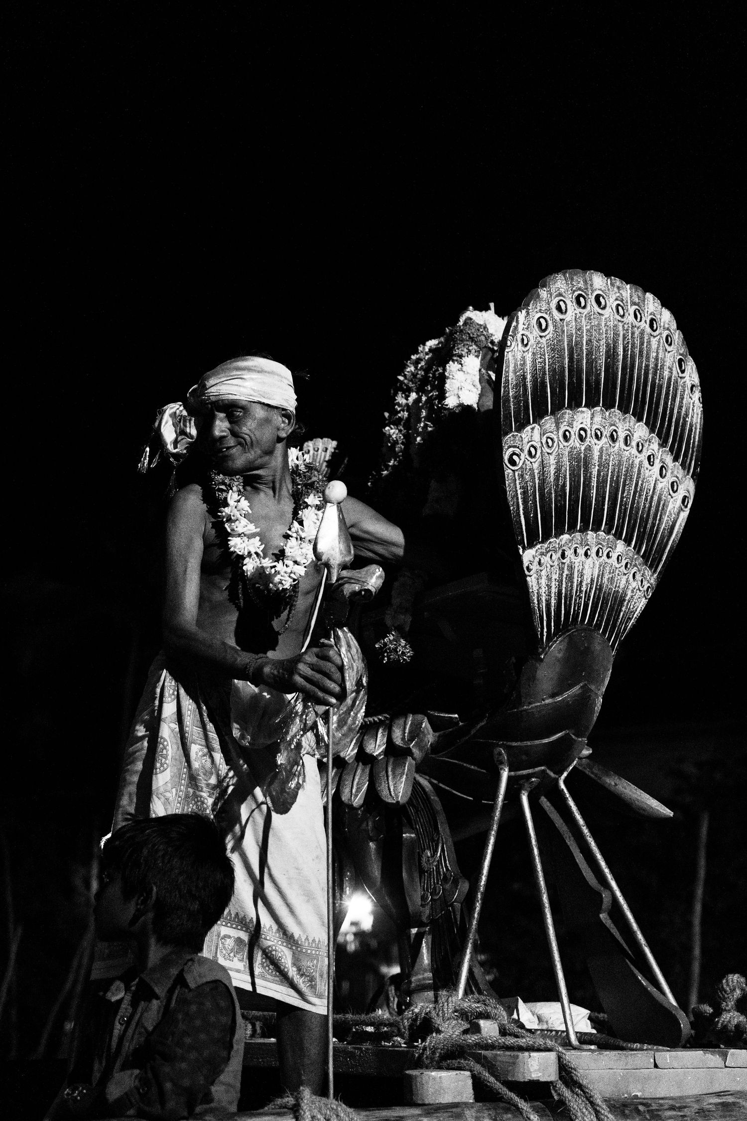 Hawaiian performer stands beside ornate feathered kahili standards during nighttime ceremony.