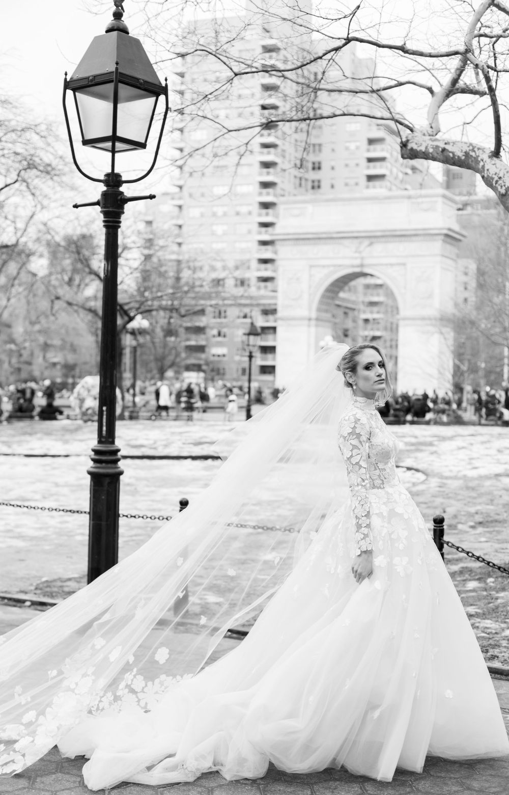 Black and white bridal photo with flowing veil and classic lamppost in urban park setting.