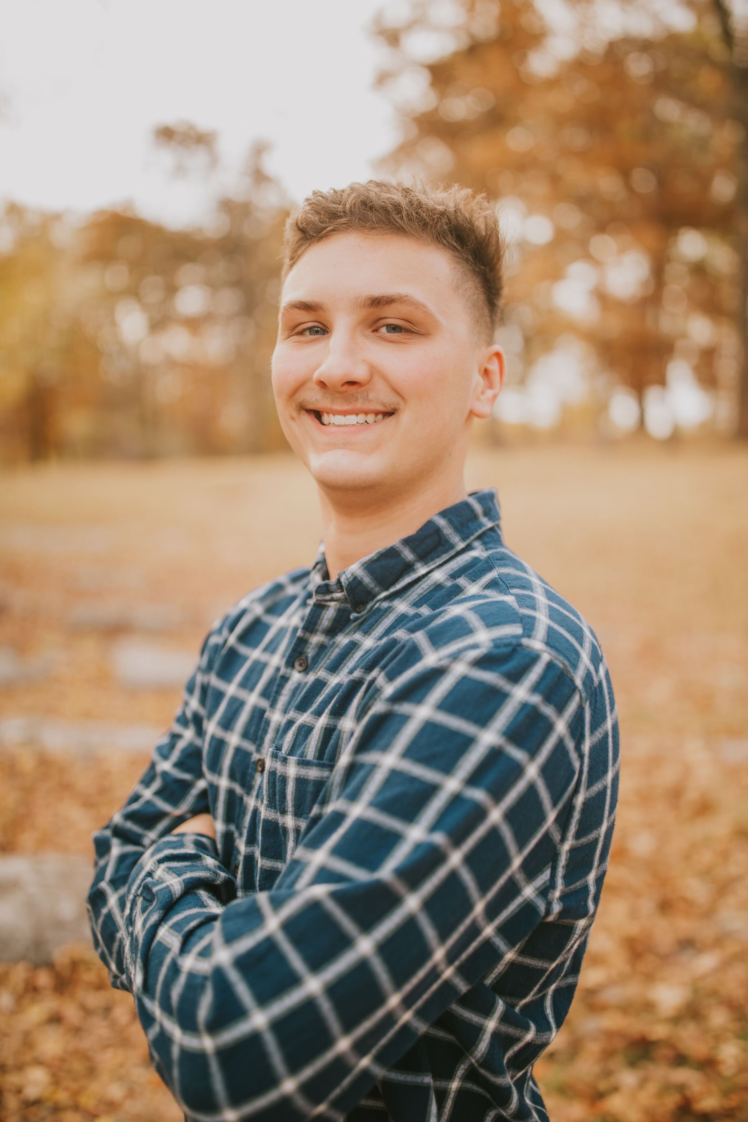 Portrait of someone wearing a plaid flannel shirt smiling in an autumn setting with golden leaves.