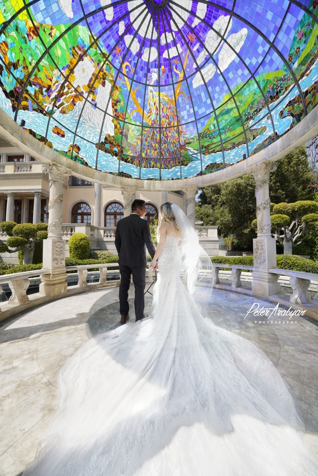 Elegant wedding couple standing under stunning stained glass dome in ornate white gazebo.