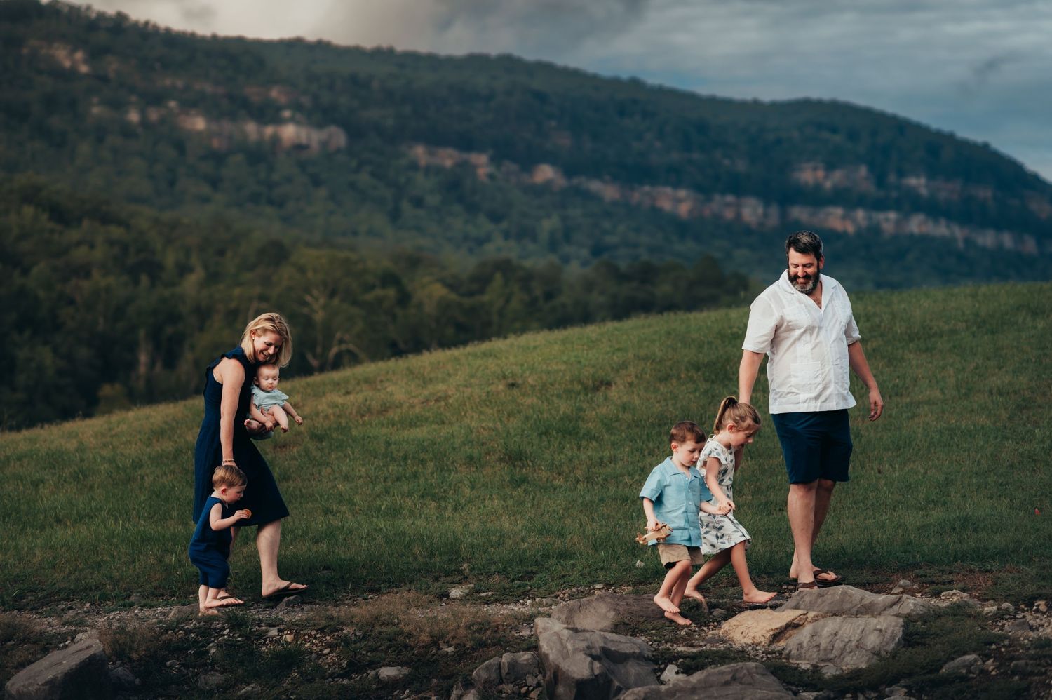 family of 6 walks in 2 groups across rocks with a mountain ridge view behind them on a sunny day