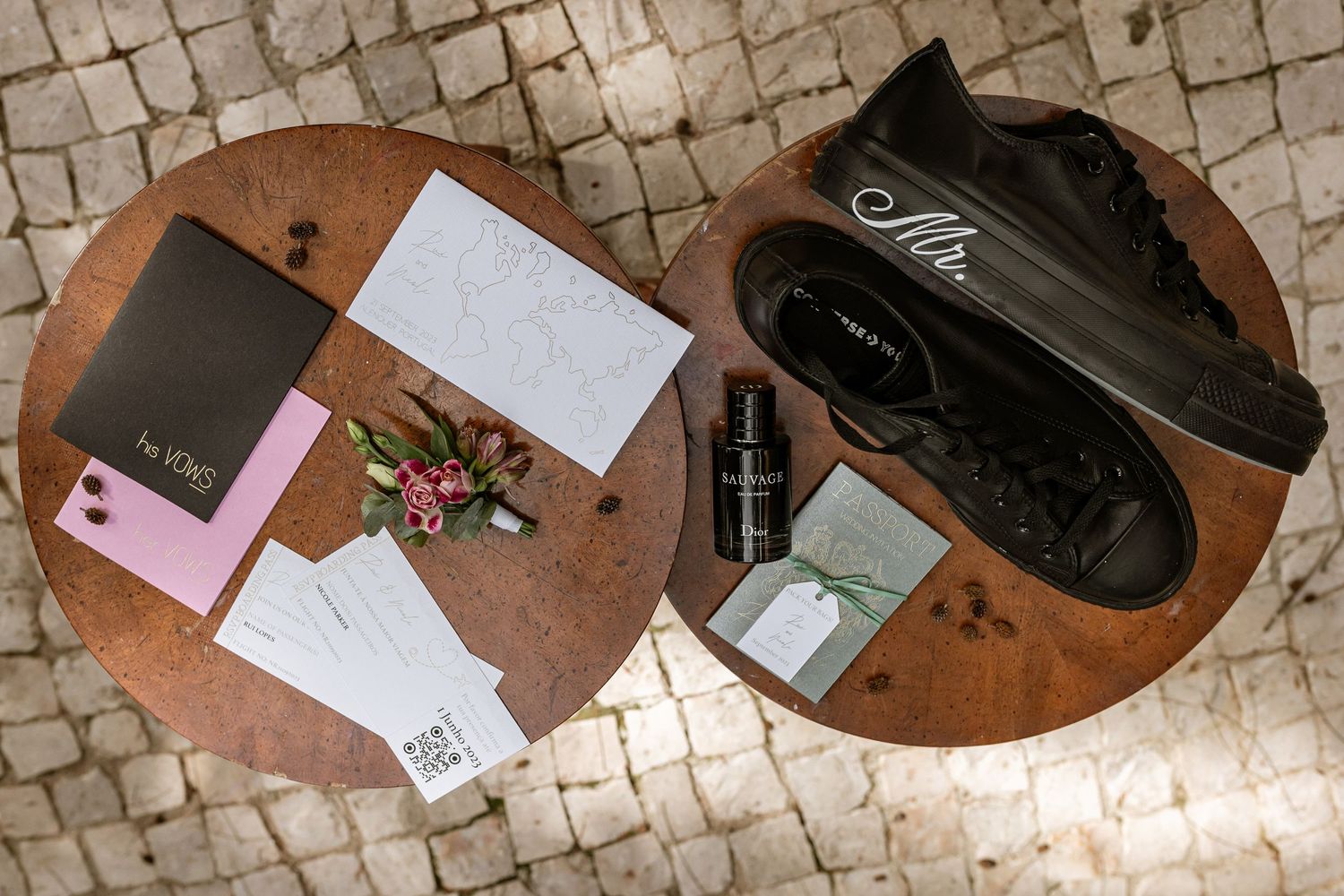 Flatlay of wedding stationery, flowers and black sneakers arranged on round wooden tables with tile floor background.