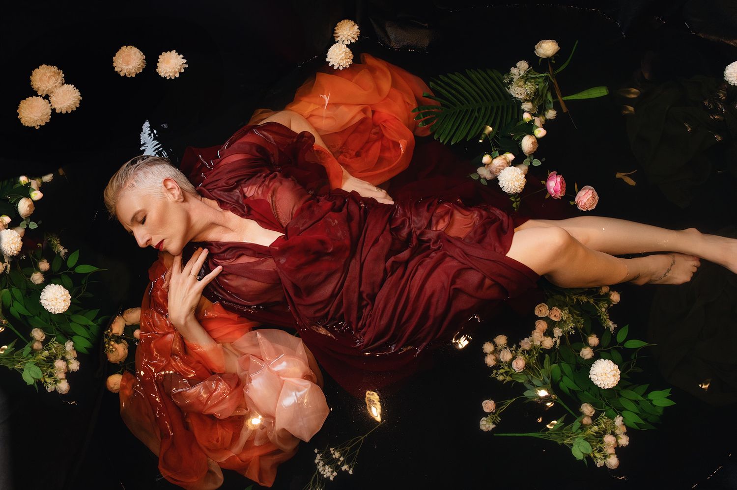 Woman in red gown floating among flowers and lights on dark water in artistic fashion photo.