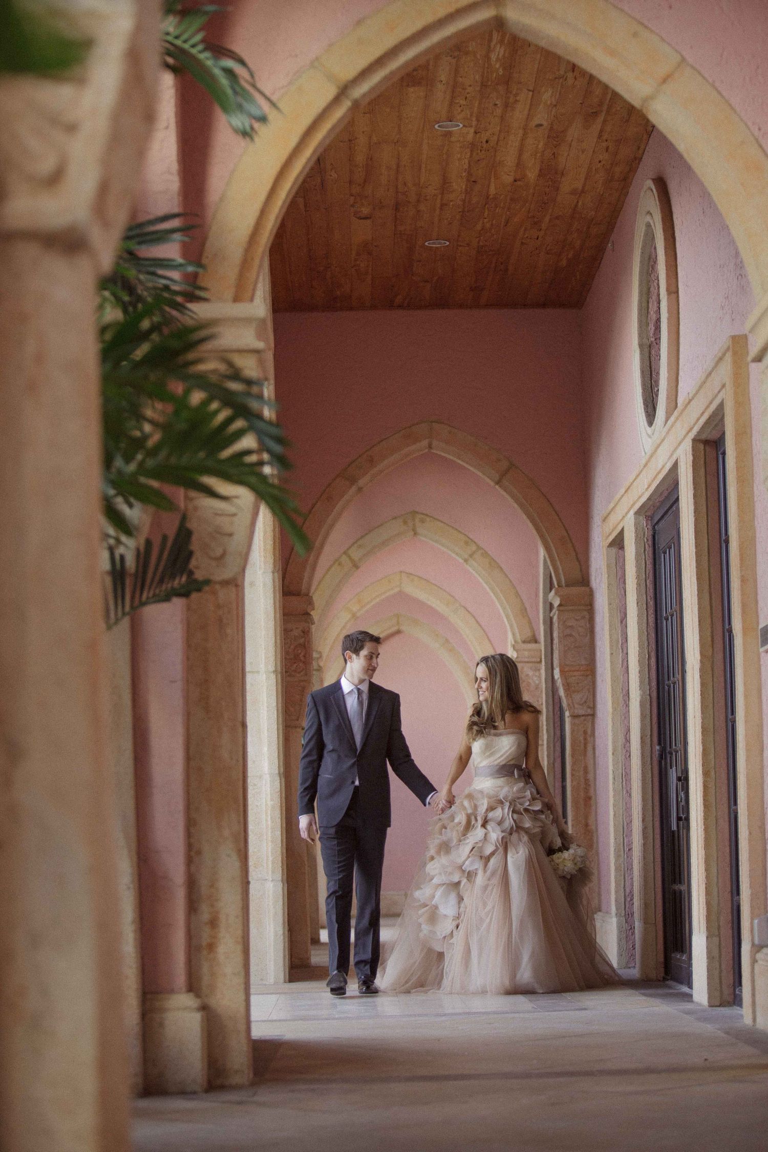 A couple in wedding attire shares an intimate moment under a pink-arched covered walkway with stone columns.