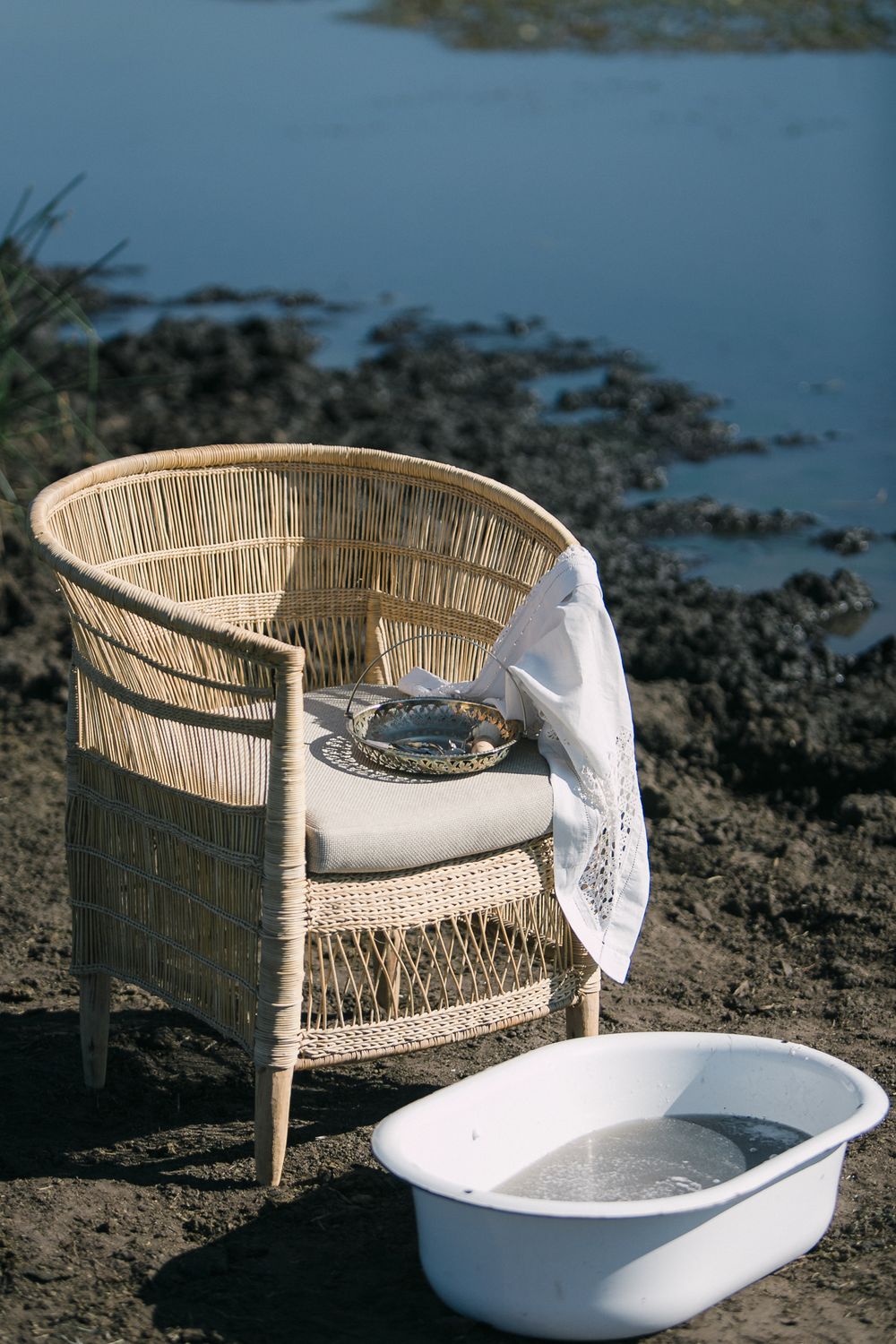 A wicker chair standing next to the blue water of a dam amongst the mud.