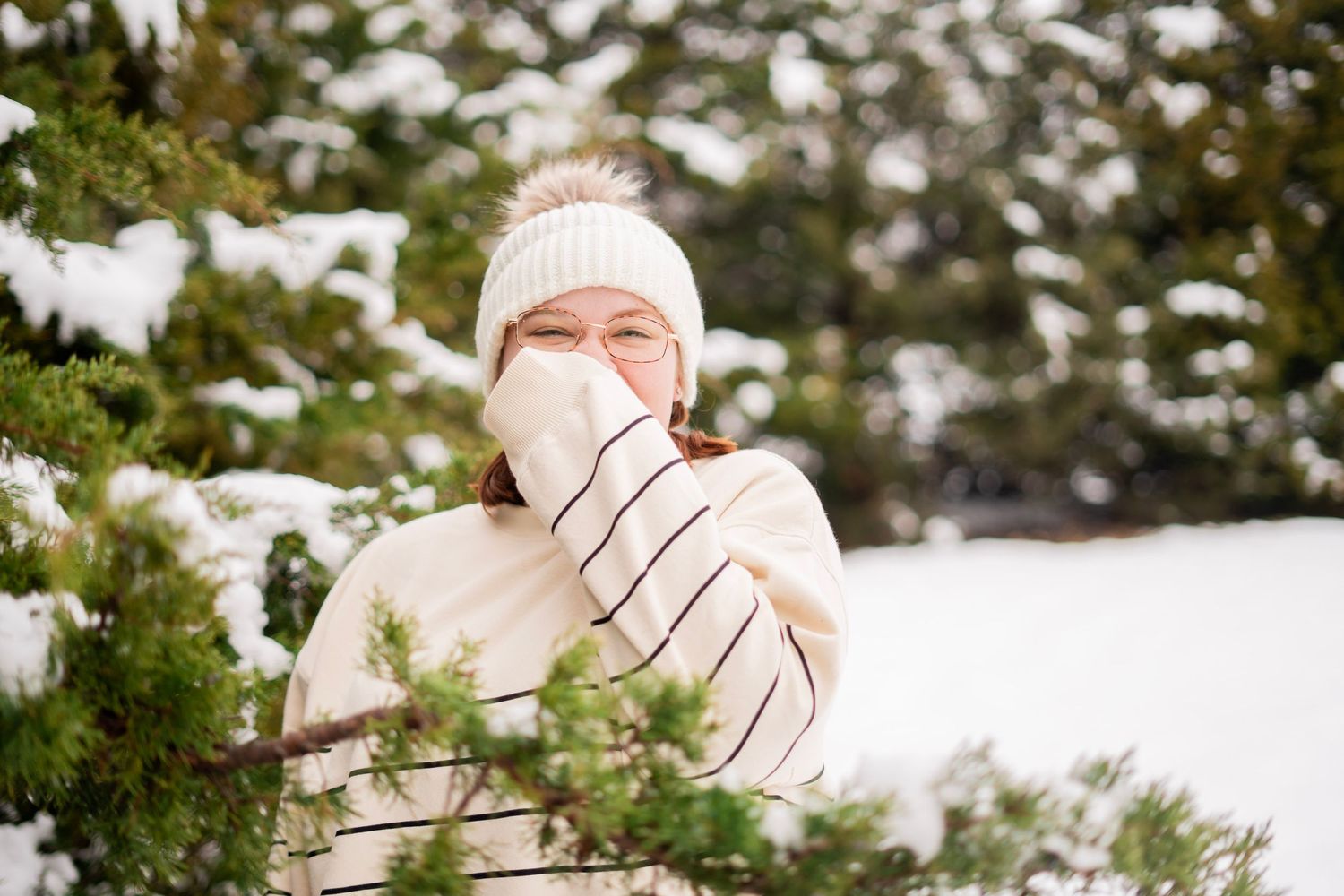 Young woman in white knit hat blowing her nose with tissue among snow-covered pine trees in winter.