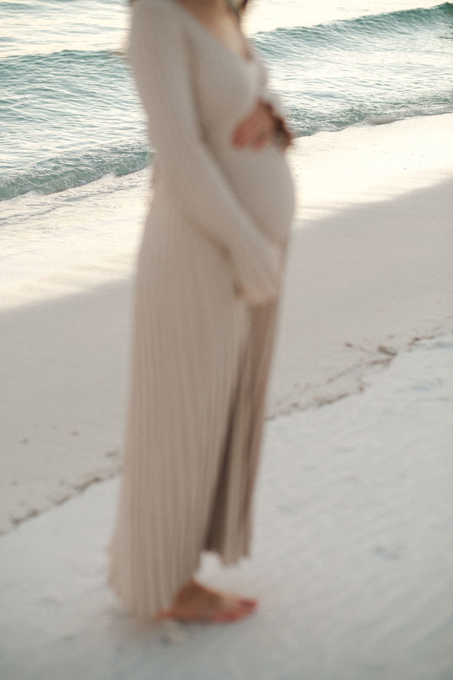 Two people in light colored clothing stand together on a sandy beach at sunset.