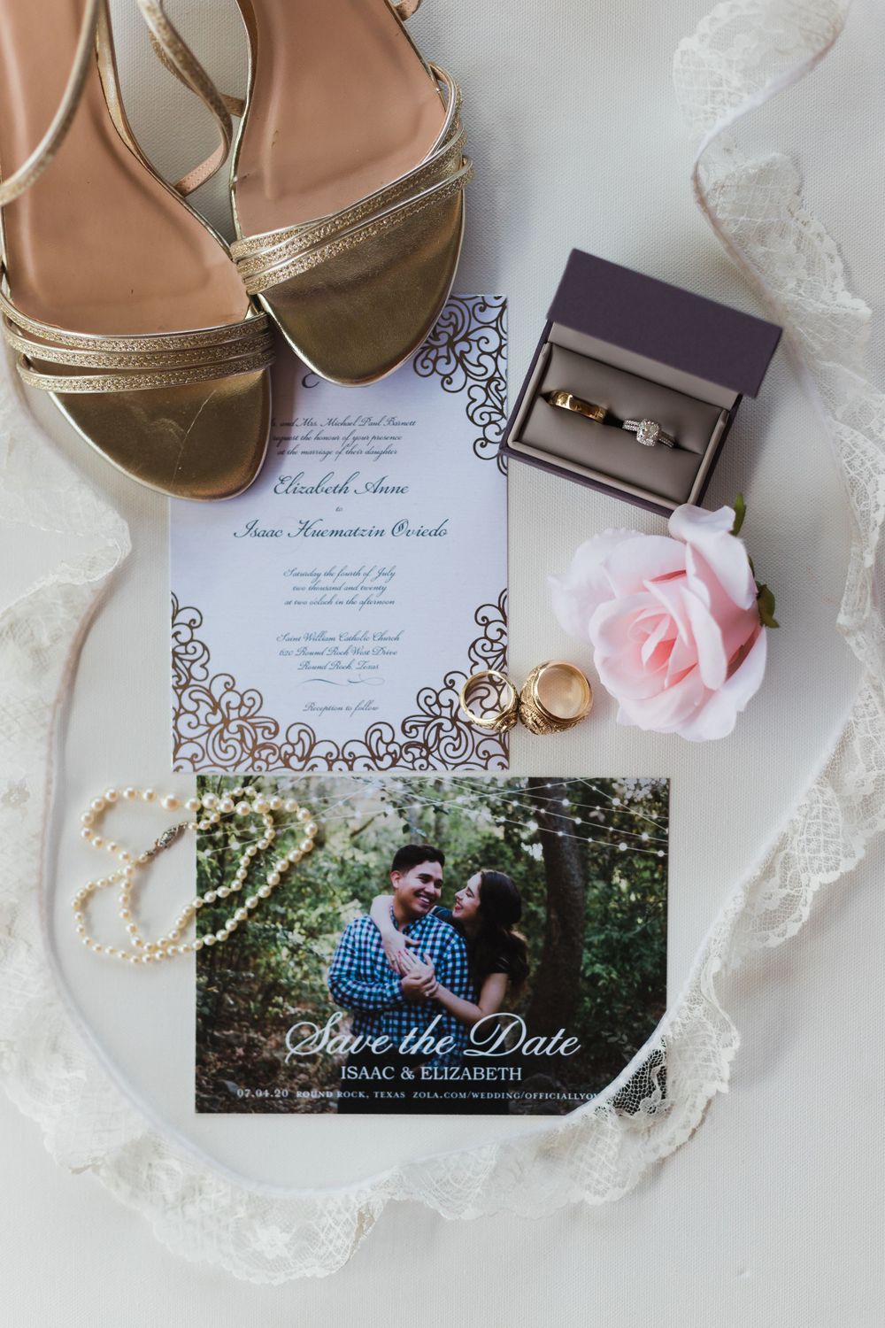 Wedding details flatlay with gold shoes, invitation, rings, and pink rose on white lace.