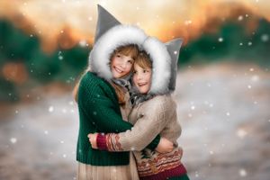 Two children in winter coats with fur-trimmed elf hoods embracing against snowy ethereal winter background.