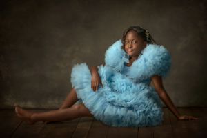 An elegant portrait featuring a young girl in a flowing blue tulle dress against a dark background.