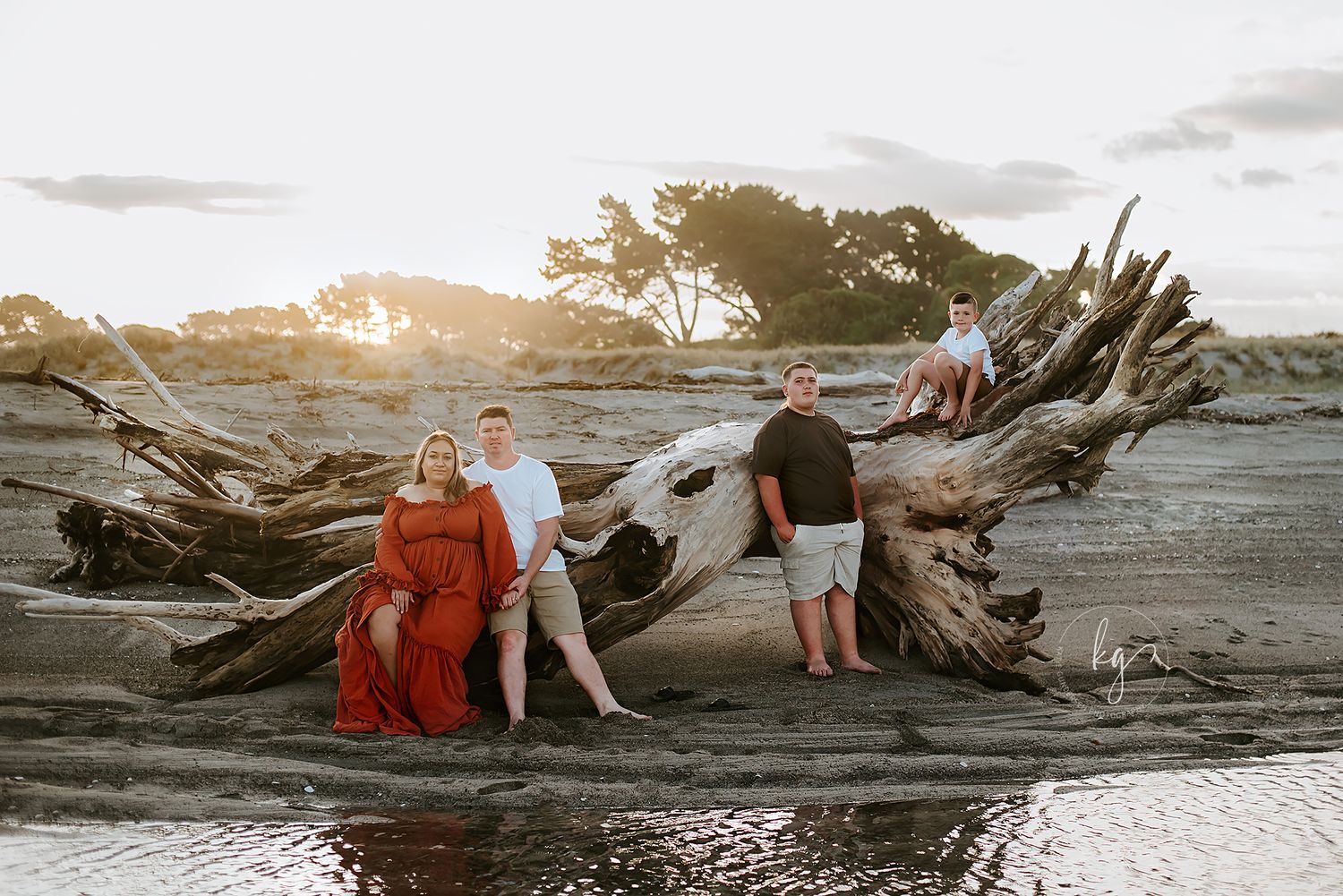 Sunset beach portrait with driftwood and red dress against golden sky.