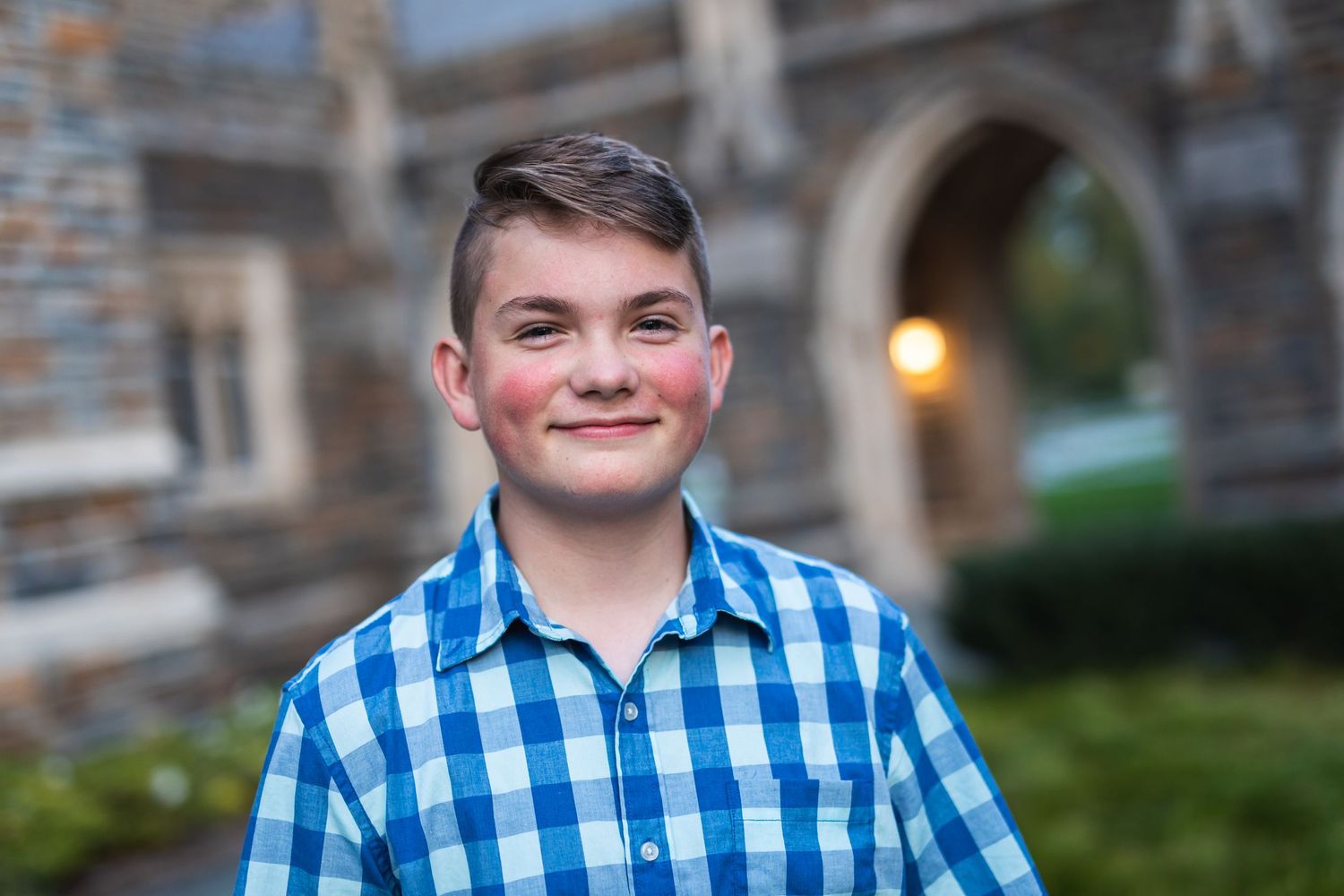 Portrait of a young person in a blue checkered shirt against a stone building backdrop.
