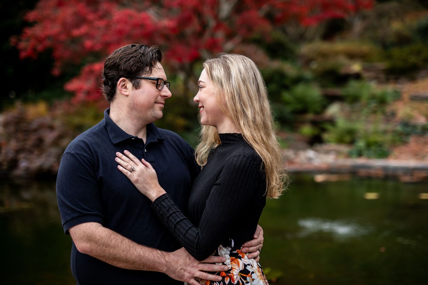A romantic couple poses by a pond with vibrant red maple trees in the background.
