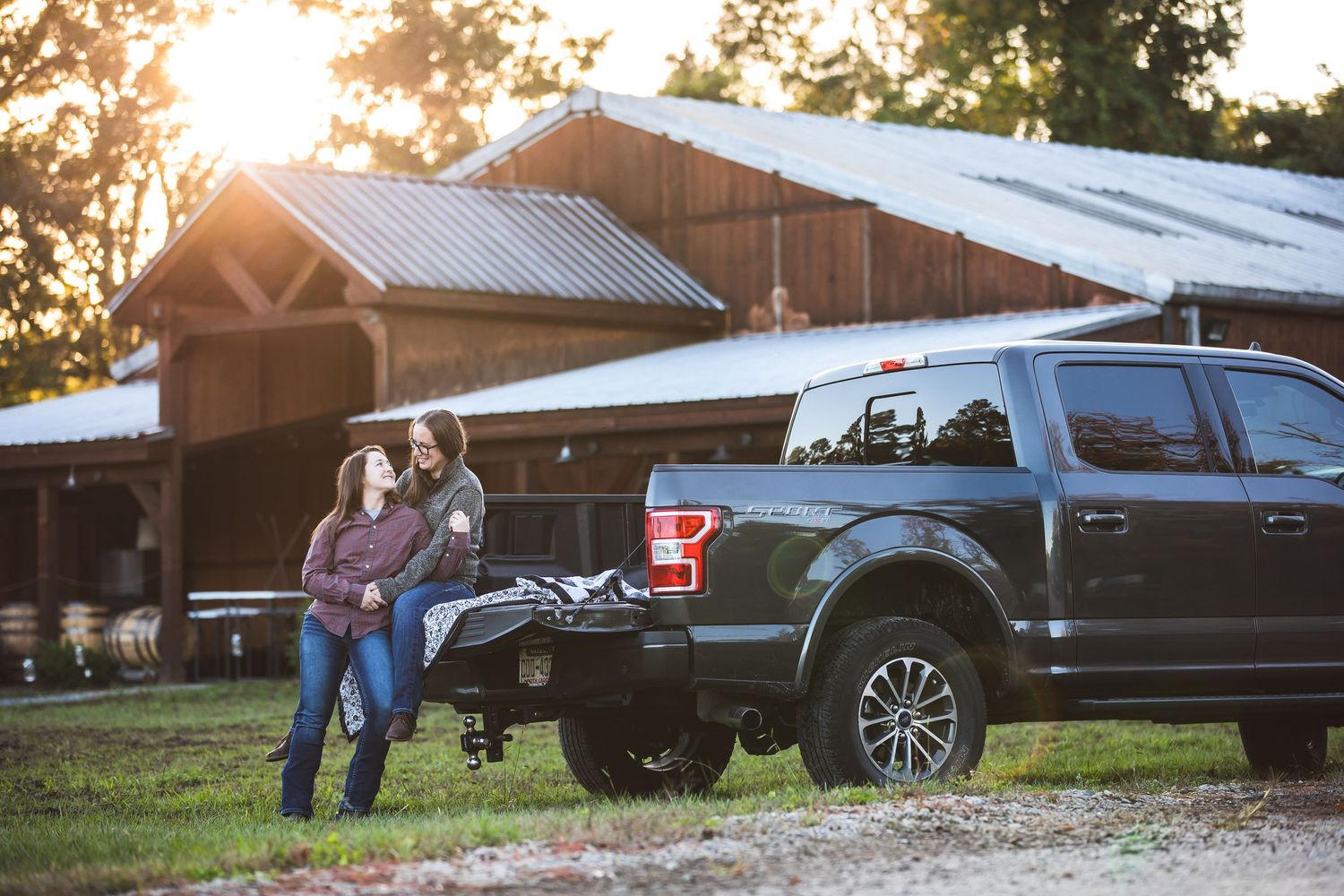 A couple stands beside a pickup truck at sunset near a rustic barn building.