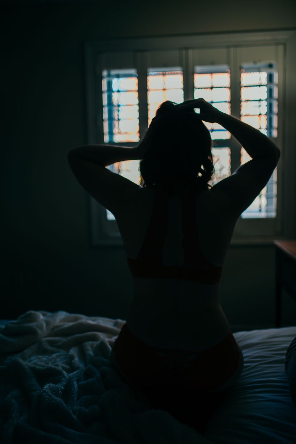 Silhouette of a woman sitting on bed against window with shutters.