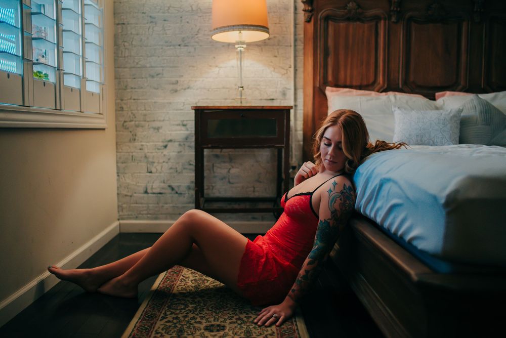 Woman in red dress poses on floor near bed in ambient room lighting.