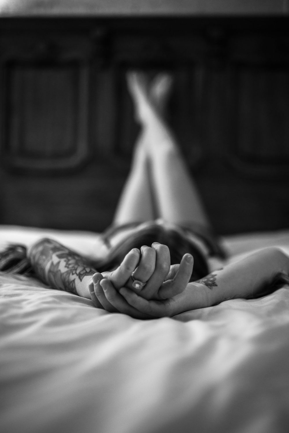 Artistic black and white photo of hands clasped on white bedding with crossed legs in background.