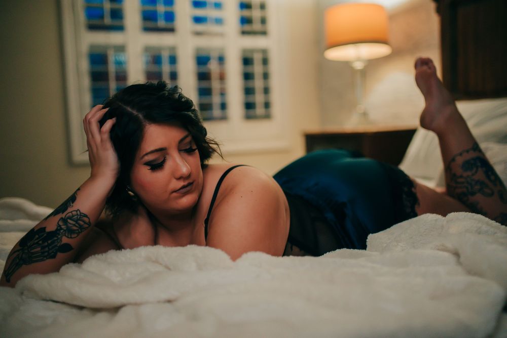 Moody boudoir portrait in teal lingerie with city windows and orange lamp.