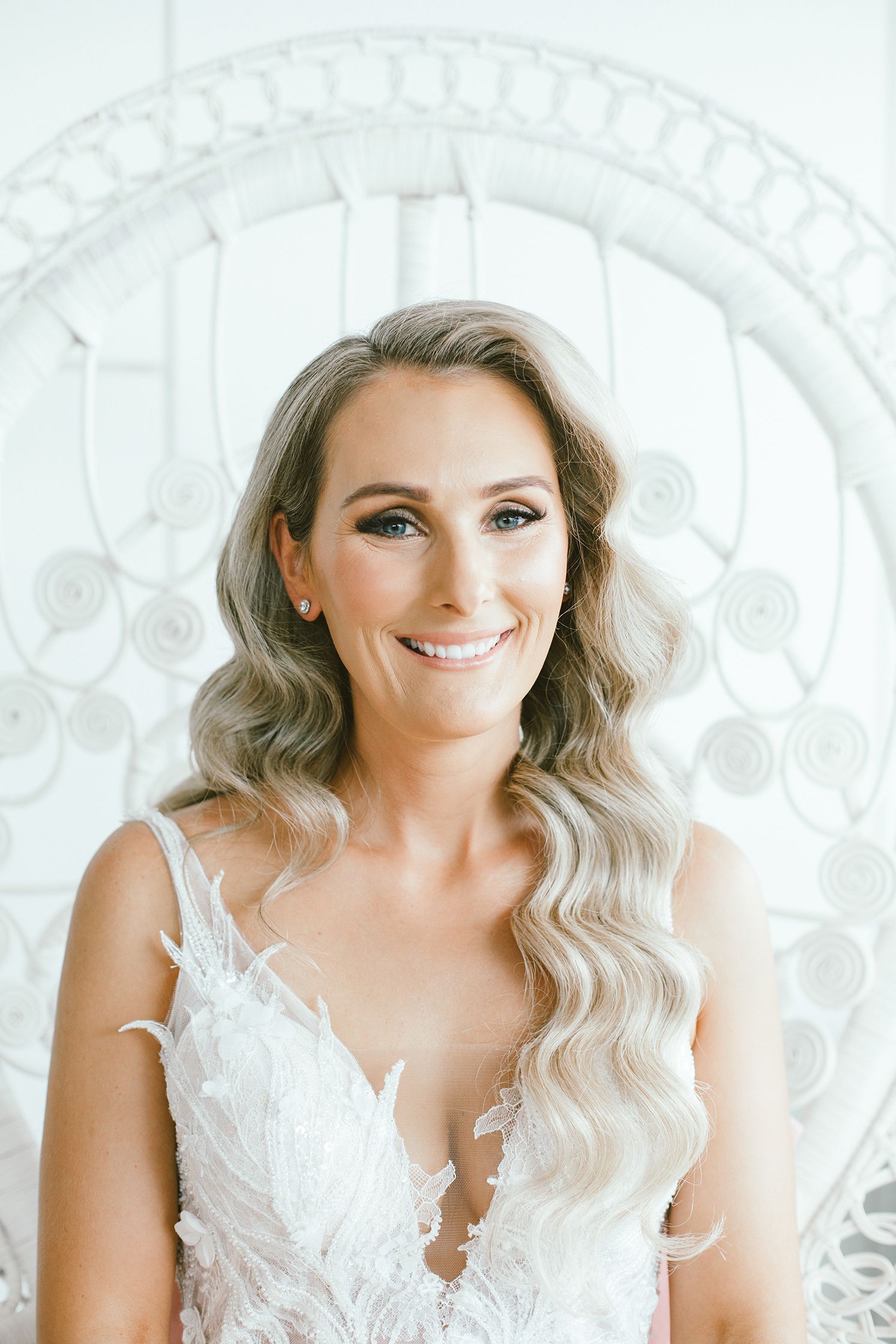 Elegant bridal portrait showcasing wavy hair and intricate lace dress against white wicker backdrop.