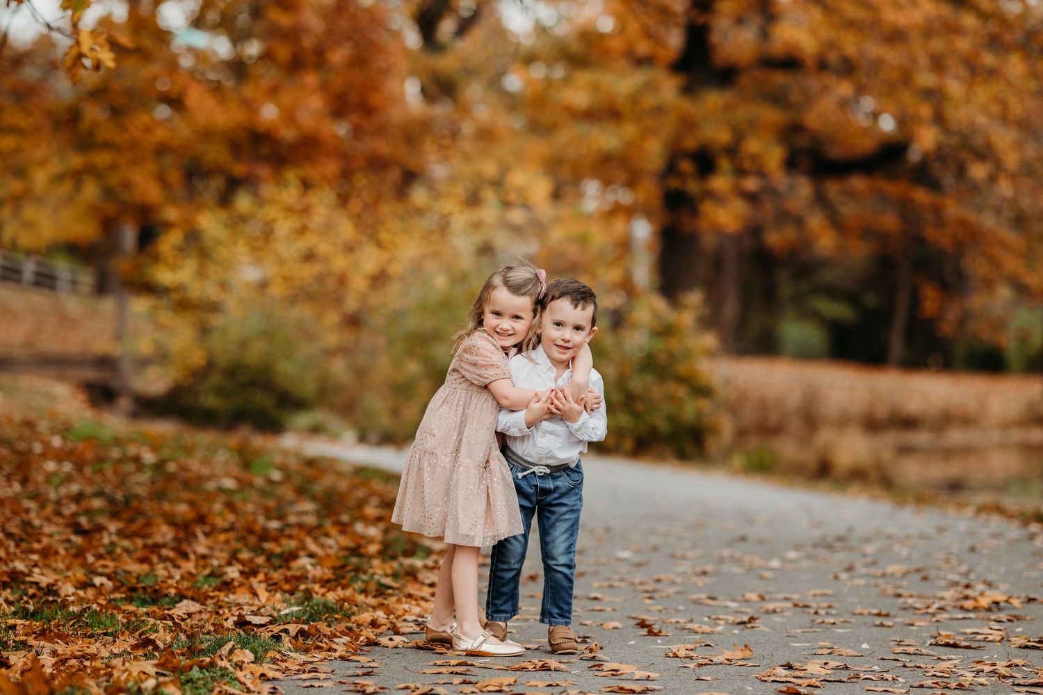 Two children embrace while standing on a path surrounded by vibrant autumn foliage and fallen leaves in Montclair.