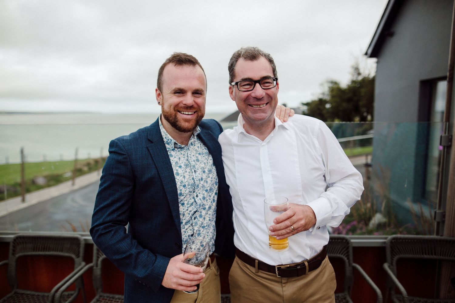 Two wedding guests share drinks and smiles on an outdoor balcony overlooking water.
