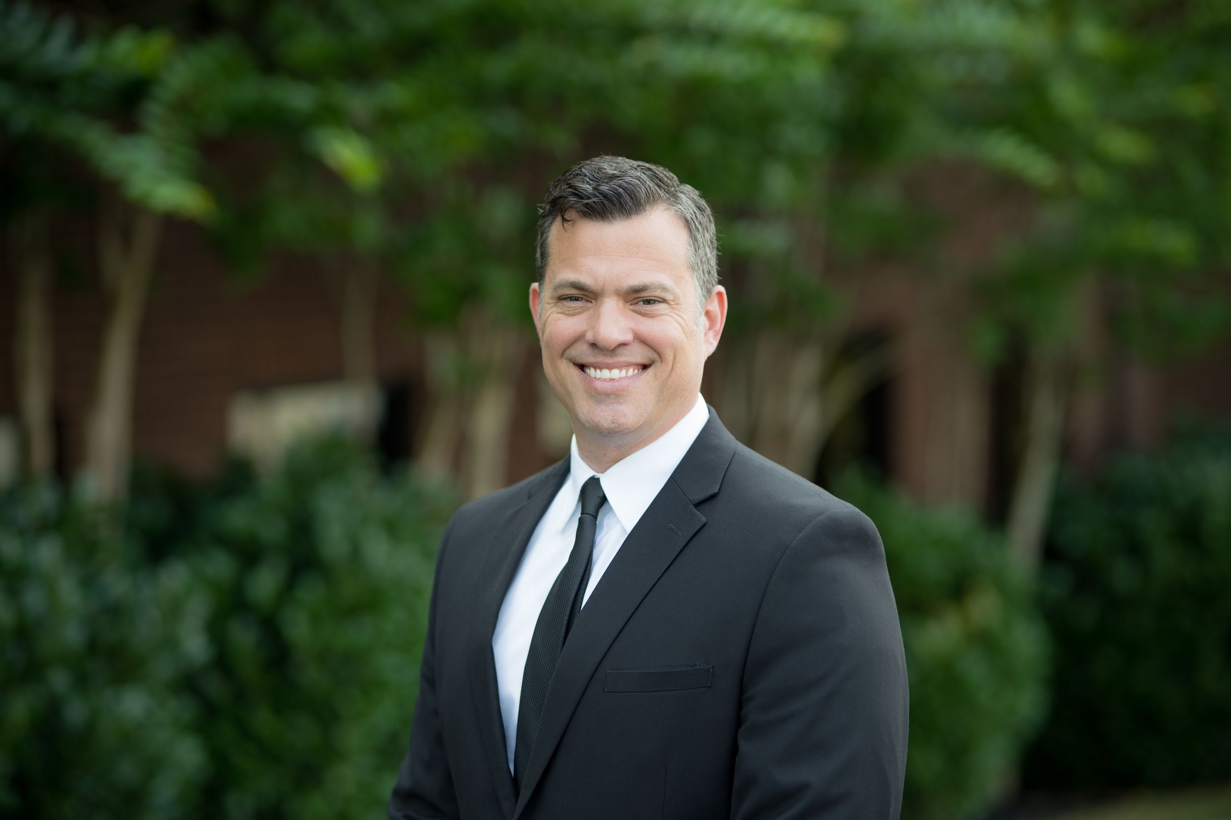 Professional headshot of person in black suit and tie smiling outdoors with greenery in background.