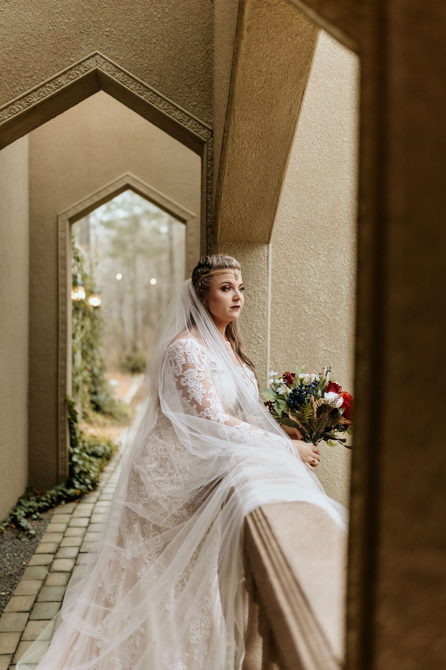 Bride in lace gown and veil poses elegantly in arched architectural passageway.