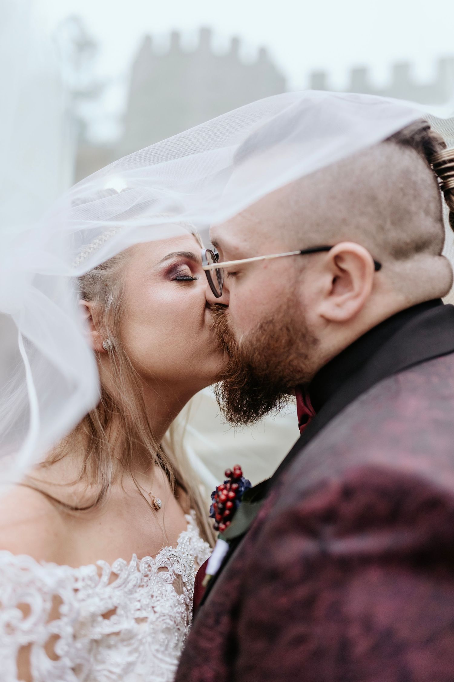 Intimate wedding moment captured with couple kissing under flowing white veil against castle backdrop.