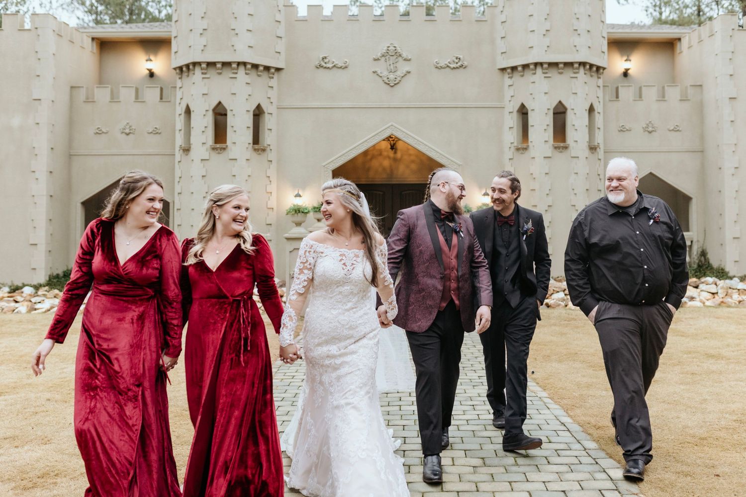 Wedding party walking and laughing together outside a castle-like venue with bridesmaids in red dresses.