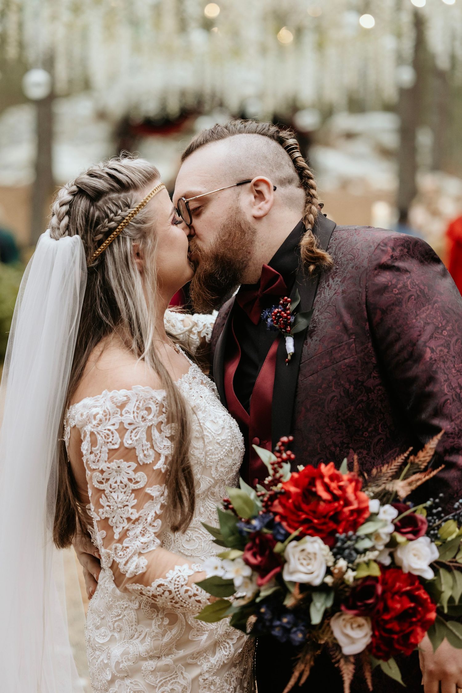 Newlyweds share intimate kiss under hanging lights with red and white floral bouquet.