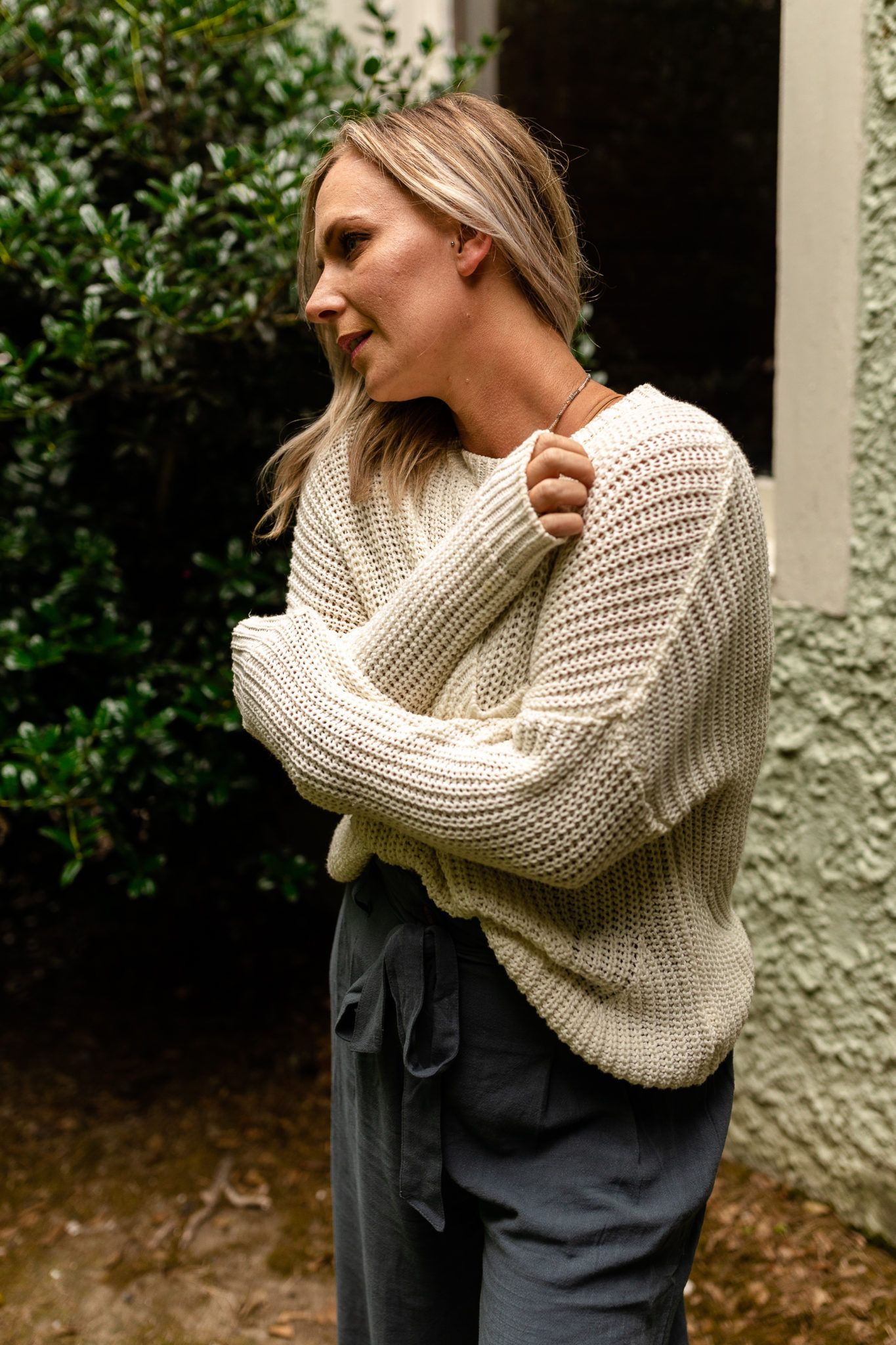 Model wearing chunky cream knit sweater and gray pants poses casually against textured wall.