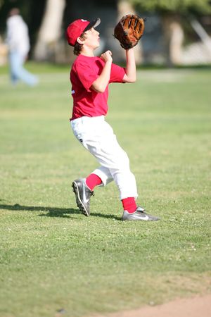 Young baseball player in red uniform reaches up to catch ball in outfield.