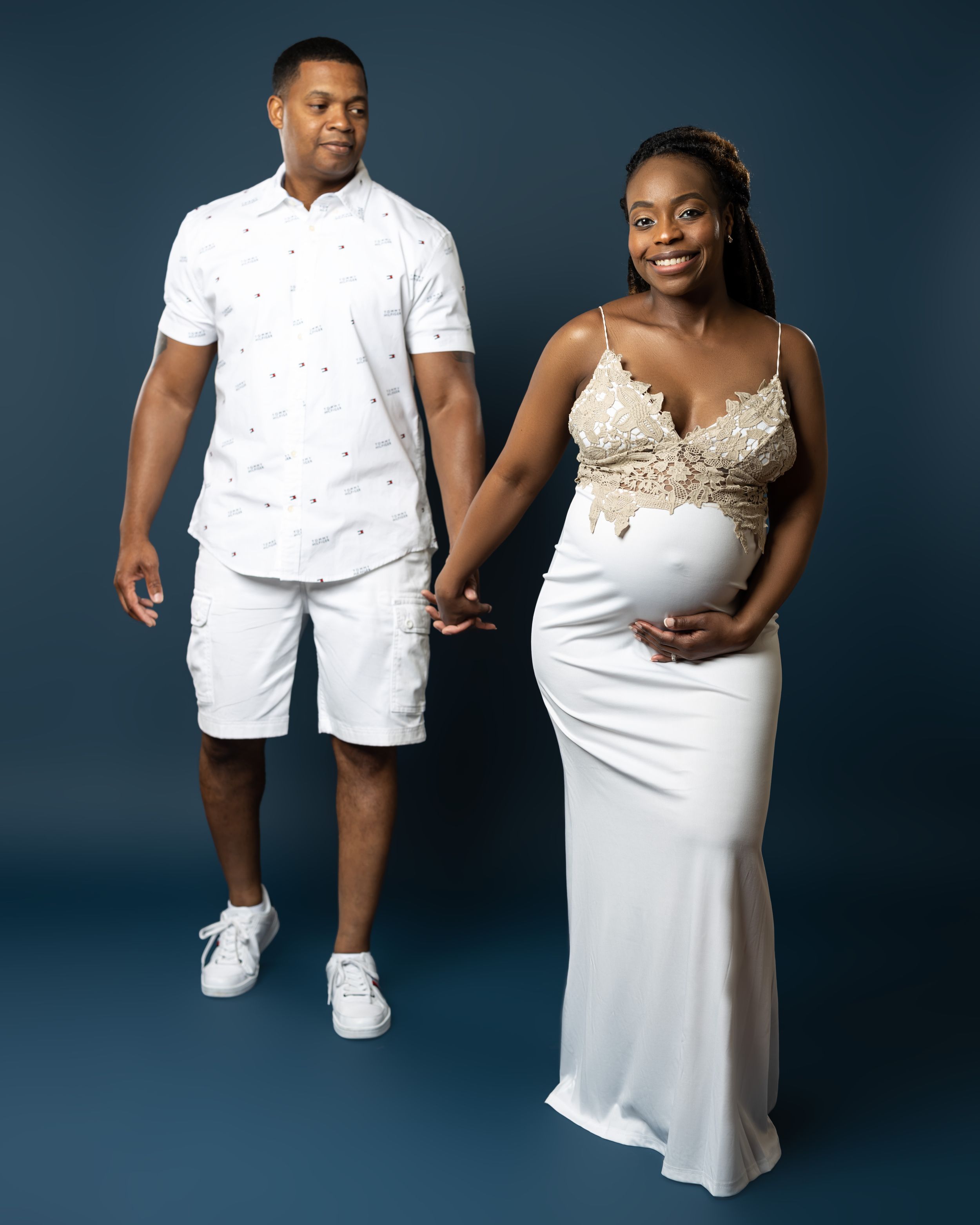 Elegant maternity portrait of a couple in matching white outfits walking against a blue background.