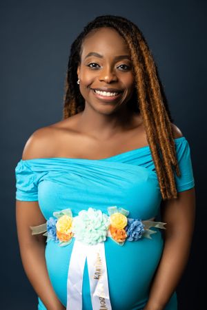 A maternity portrait in a turquoise dress with floral sash against dark background.
