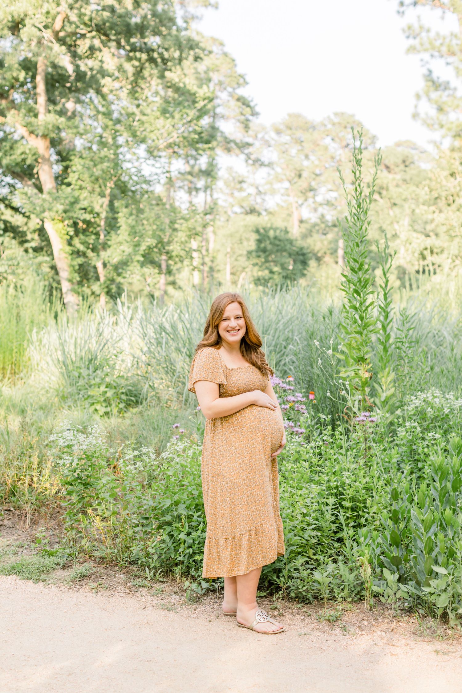 A person in tan dress poses along a garden path surrounded by tall wildflowers and trees.