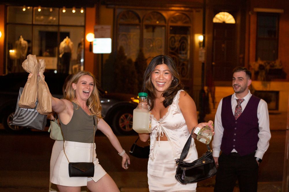 Two women laughing and carrying bags outside a restaurant at night with a man in background.