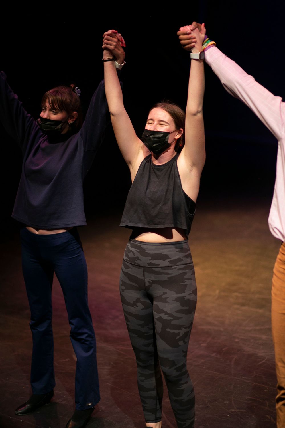 Three performers wearing black face masks take a bow holding hands on a dark stage.
