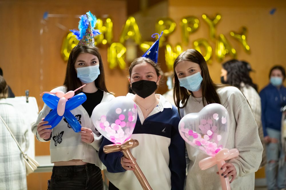 Three young women wearing masks hold balloon wands at a birthday celebration indoors.