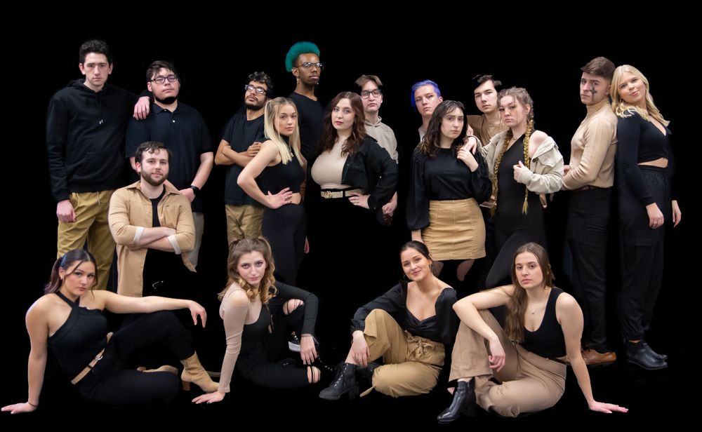A diverse group of young performers pose together in black and tan outfits against a dark background.