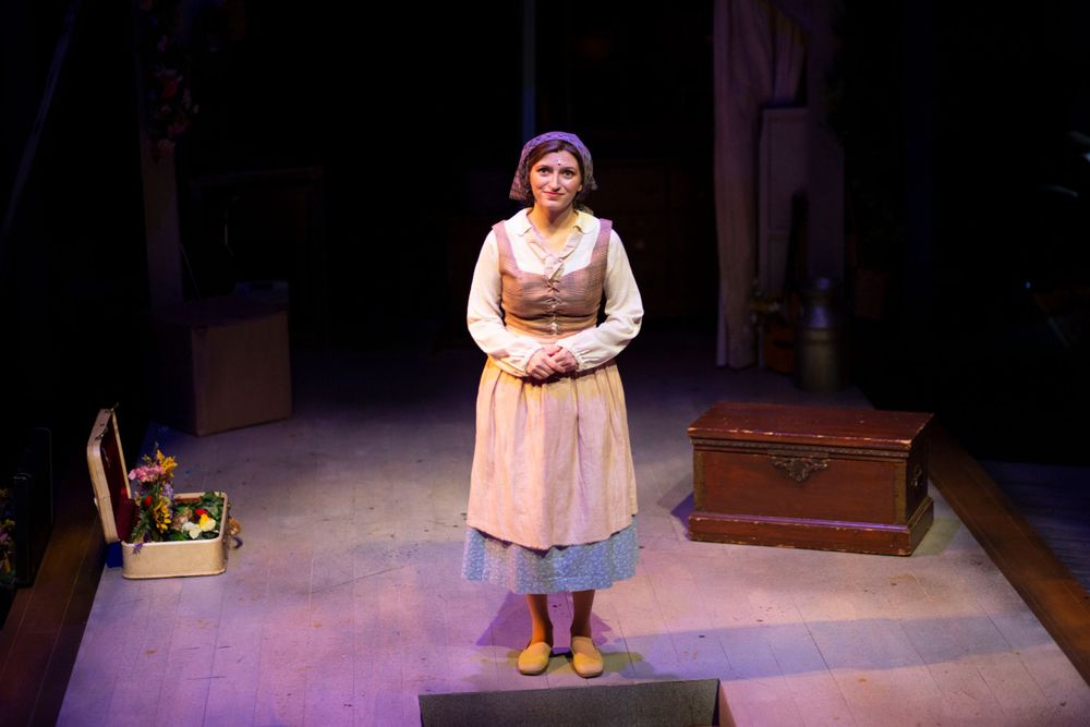 Female performer in period costume stands on stage during a theatrical production.