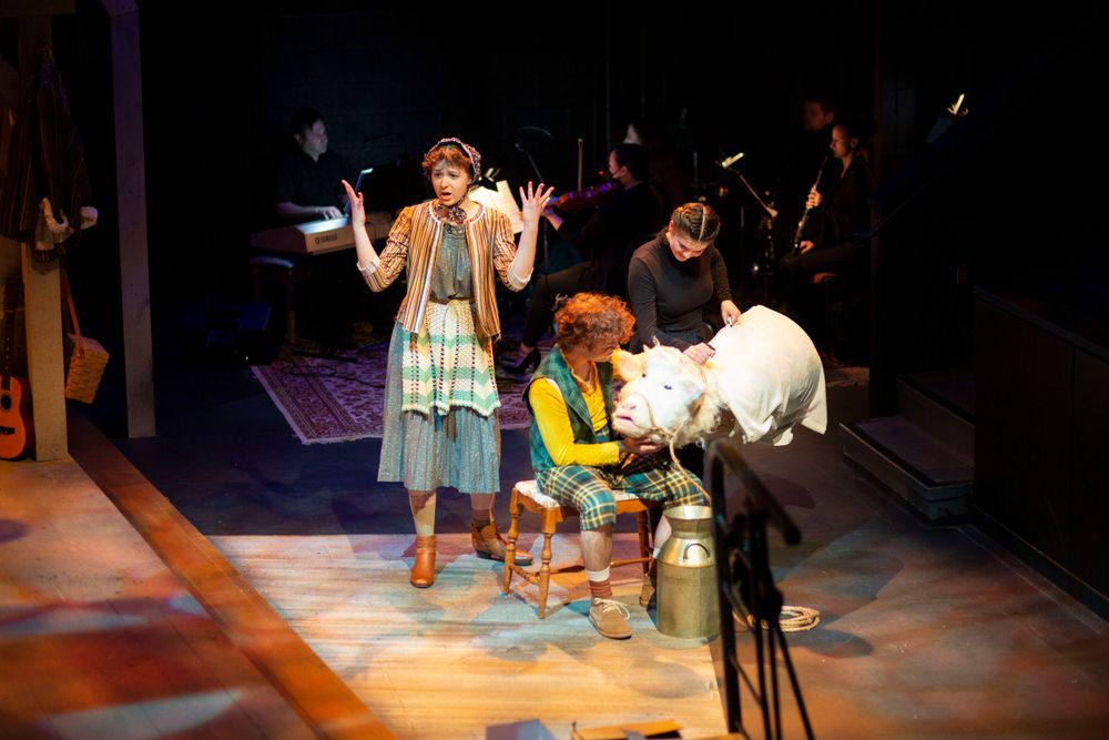 Two performers on stage during a theatrical production with musicians visible in the background.
