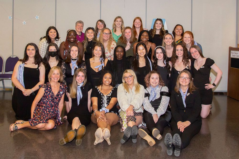Large group of women in black attire pose together for a formal group photo indoors.