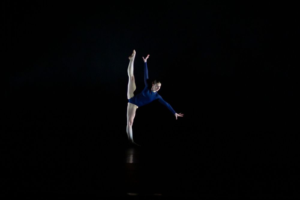 Dancer in blue leotard performs a dramatic leap against a dark stage background.