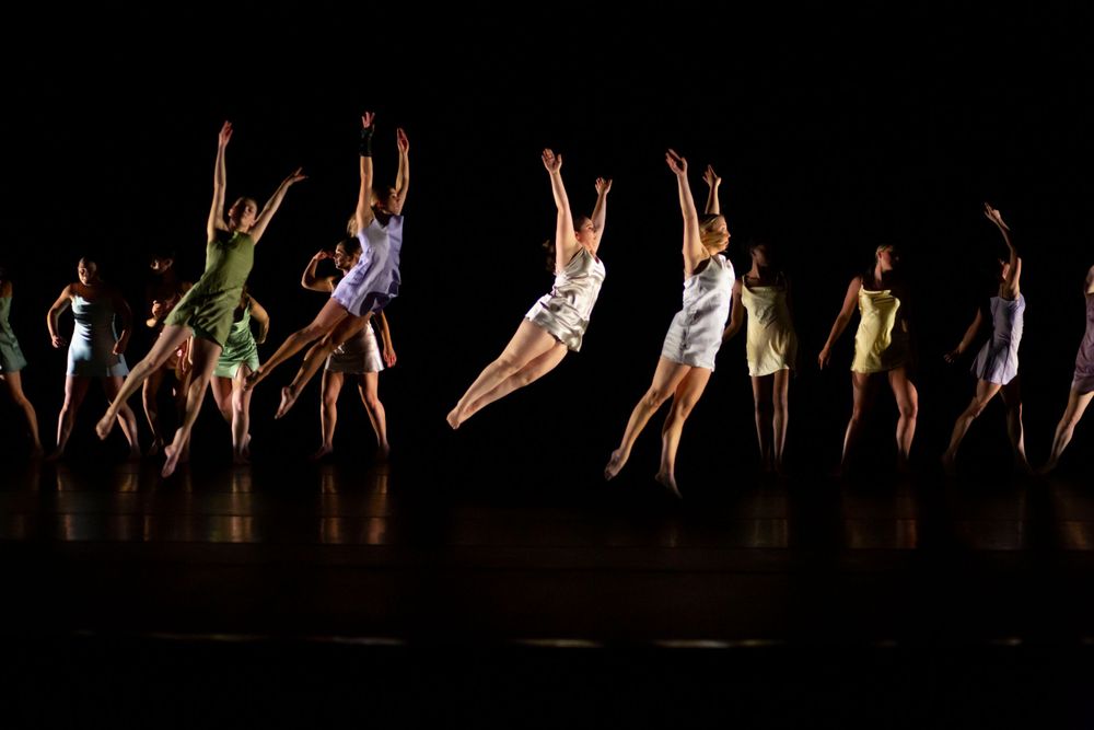 Group of dancers perform synchronized leaps on a dark stage during a dance recital.