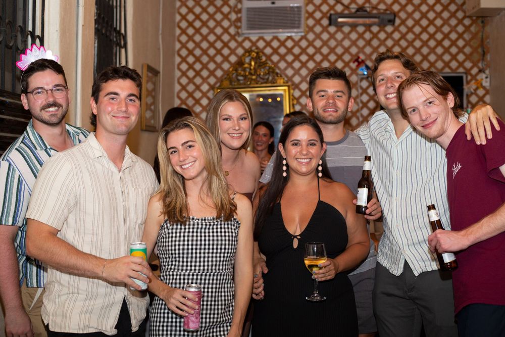 Group of young adults smiling and posing together at an indoor party, some holding drinks.