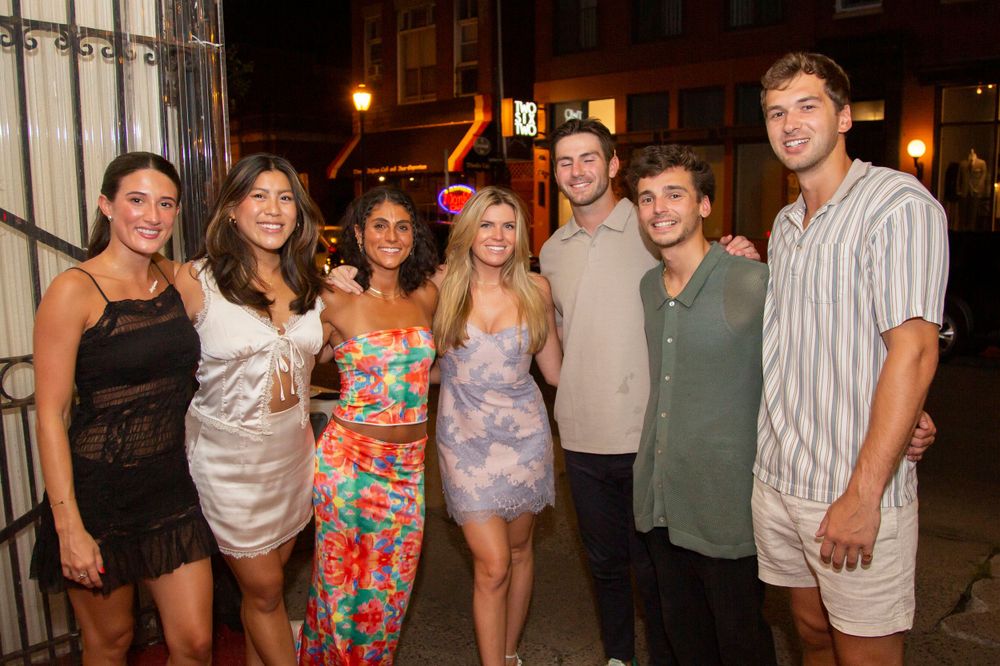 Group of seven young adults posing together outdoors at night on a city street.