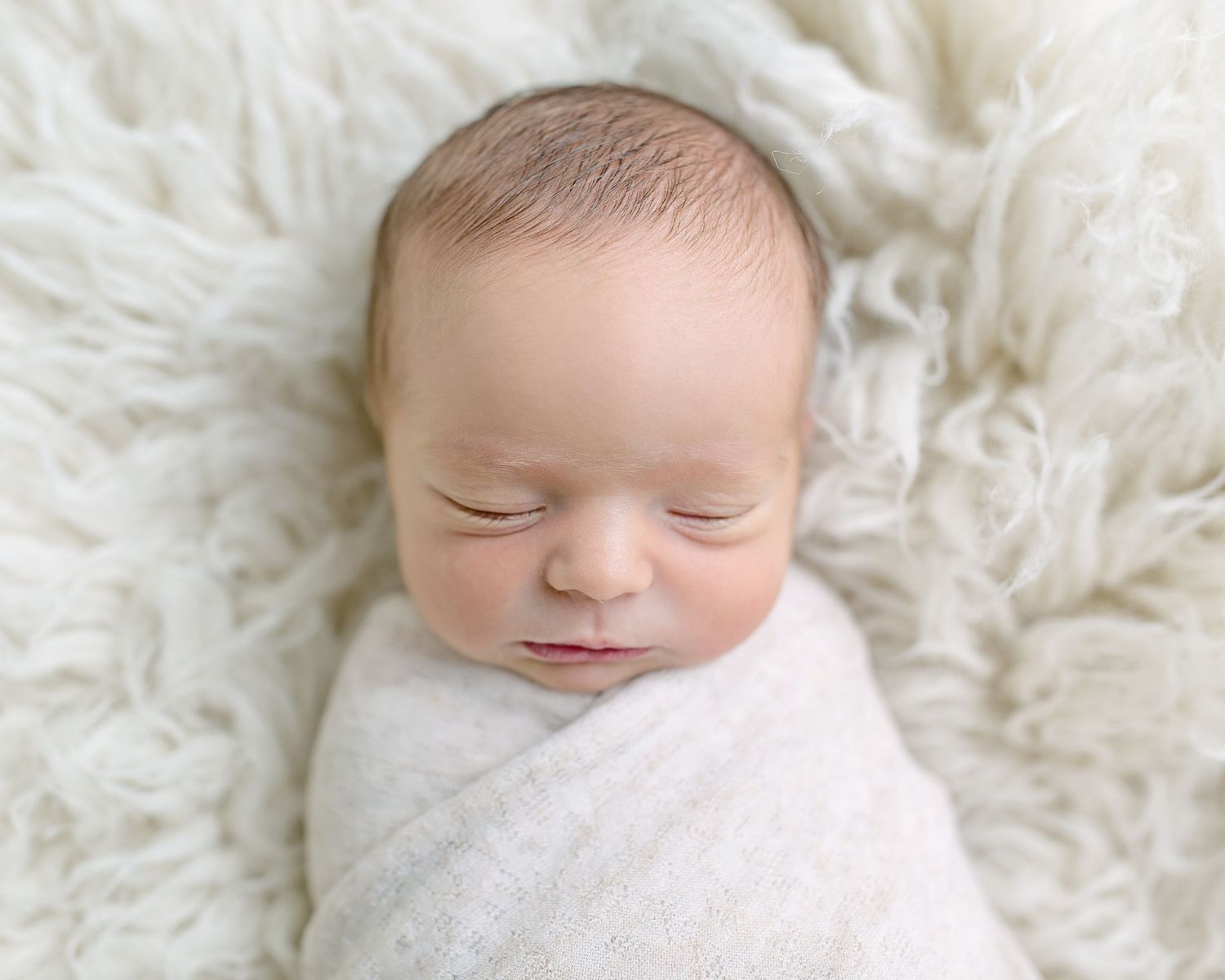 Sleeping newborn wrapped in white swaddle on fluffy white fur backdrop.