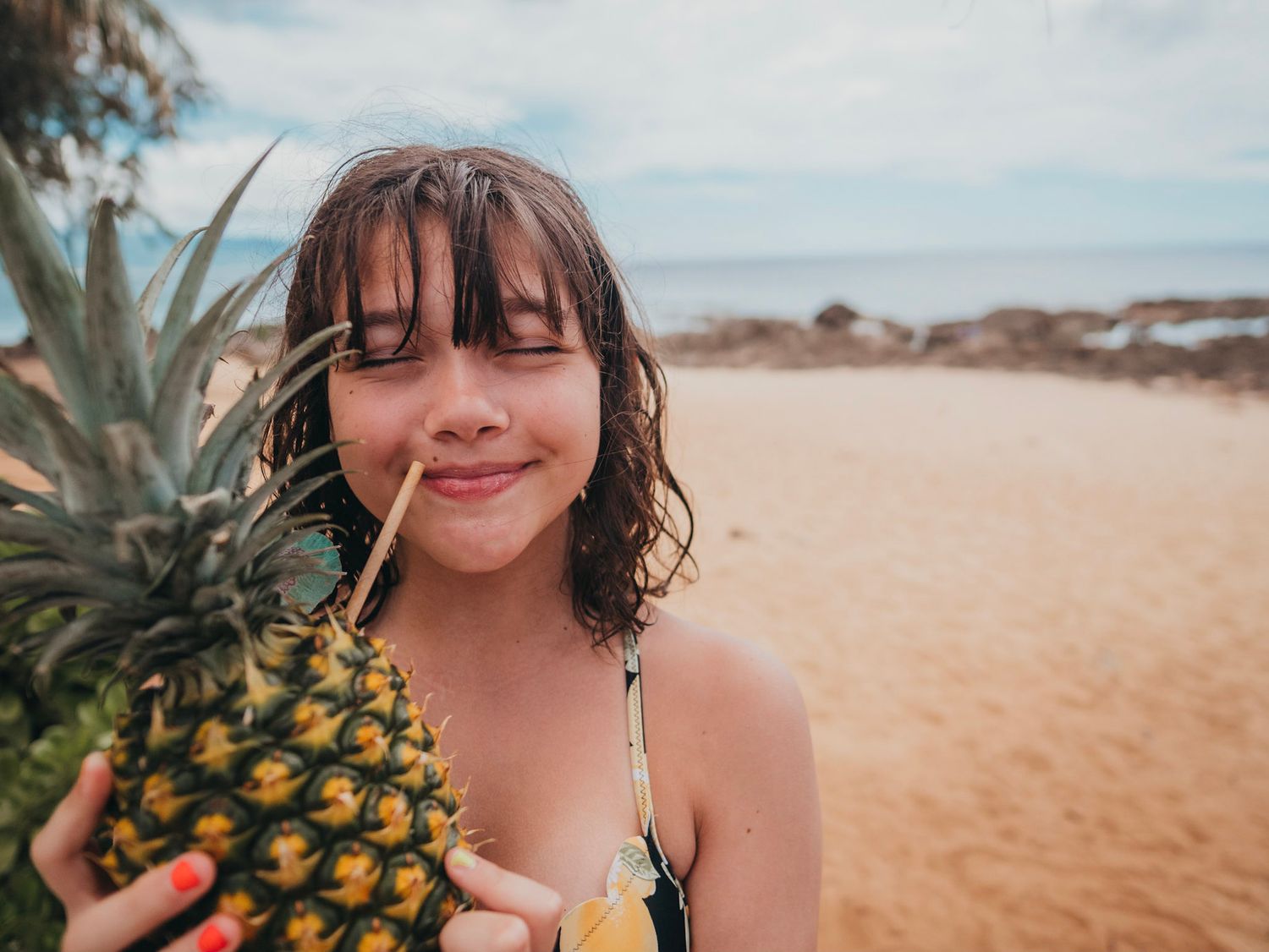 A girl on the beach in O'ahu Hawaii smiling and sipping on a pineapple cocktail by the ocean.